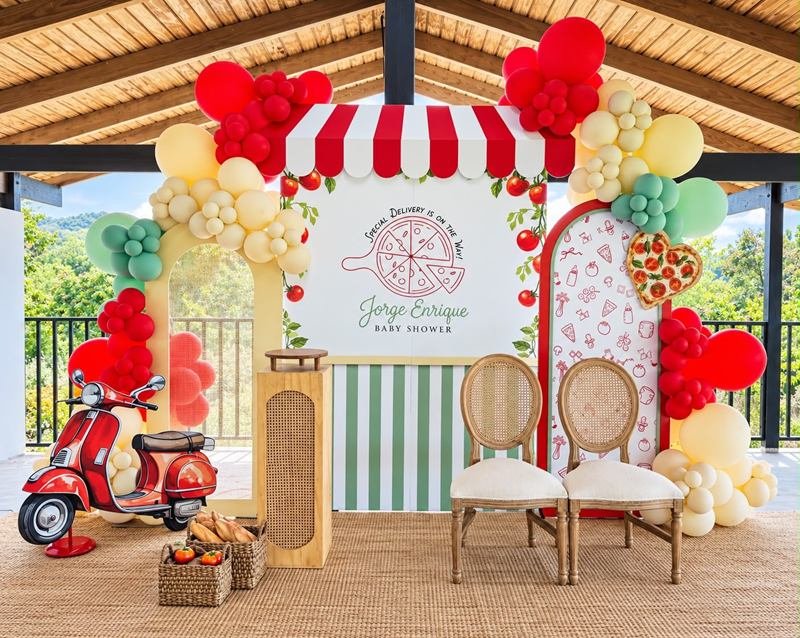 Decoración de baby shower con temática de pizzería: toldo rojo y blanco, globos rojos, crema y verdes, una motora roja decorativa y dos sillas en el área techada